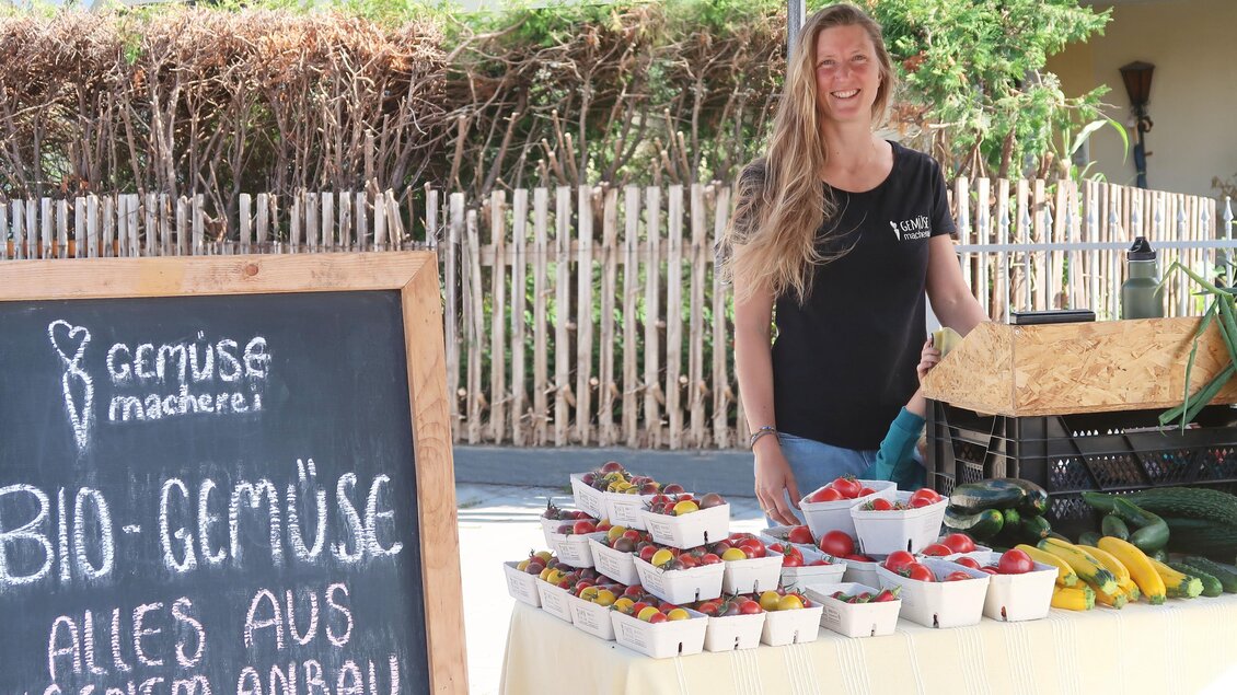 Eine Frau steht an einem Stand mit frischem bio Gemüse und Obst. Im Hintergrund ist eine Holzwand und Sonnenlicht zu sehen. | © Doll