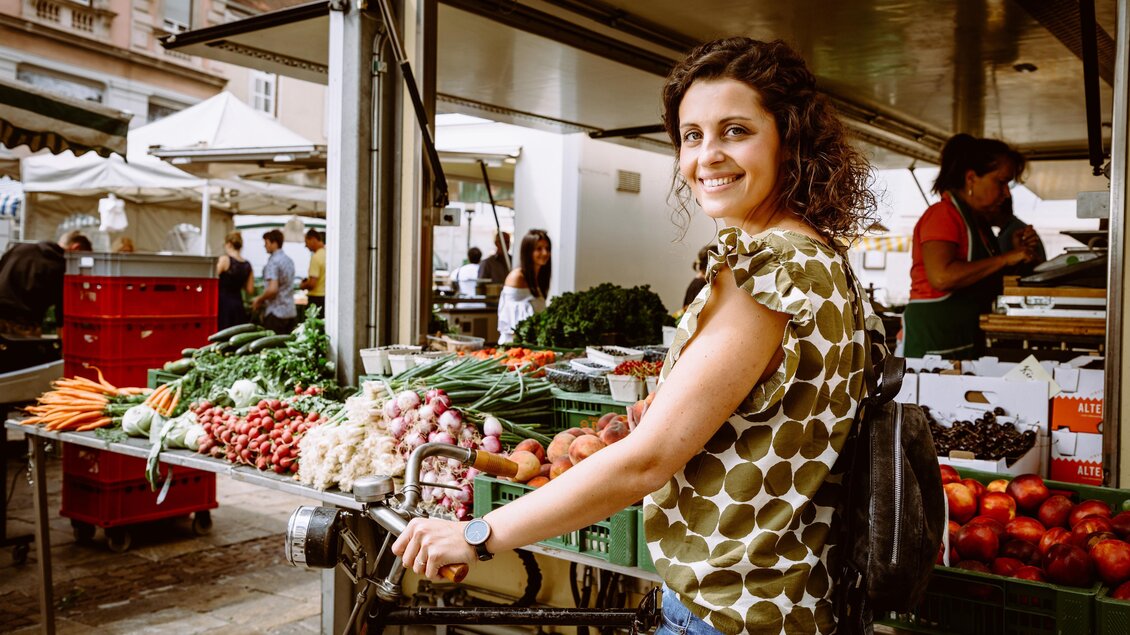 Eine Frau steht an einem Marktstand mit frischem Obst und Gemüse. Sie lächelt und trägt ein Oberteil mit Punkten. | © TVB Erzberg-Leoben