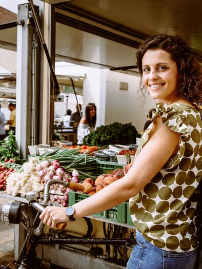 A woman is standing at a market stall with fresh fruits and vegetables. She is smiling and wearing a top with polka dots. | © TVB Erzberg-Leoben