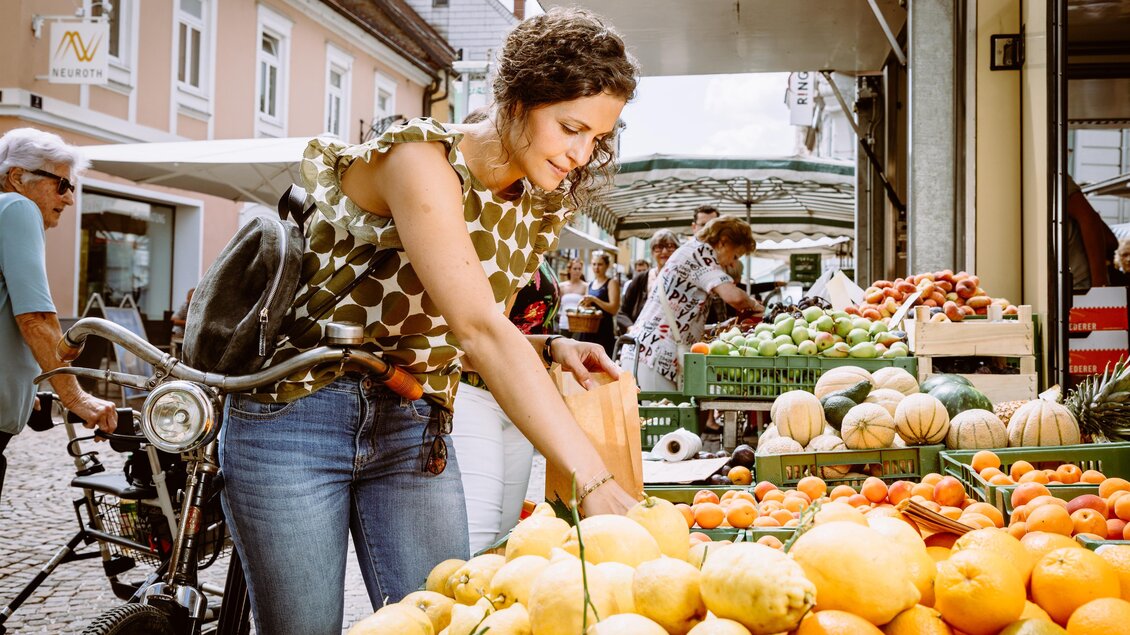 Eine Frau wählt frisches Obst auf einem belebten Markt. Im Hintergrund sind weitere Marktstände und Kunden zu sehen. | © TVB Erzberg-Leoben