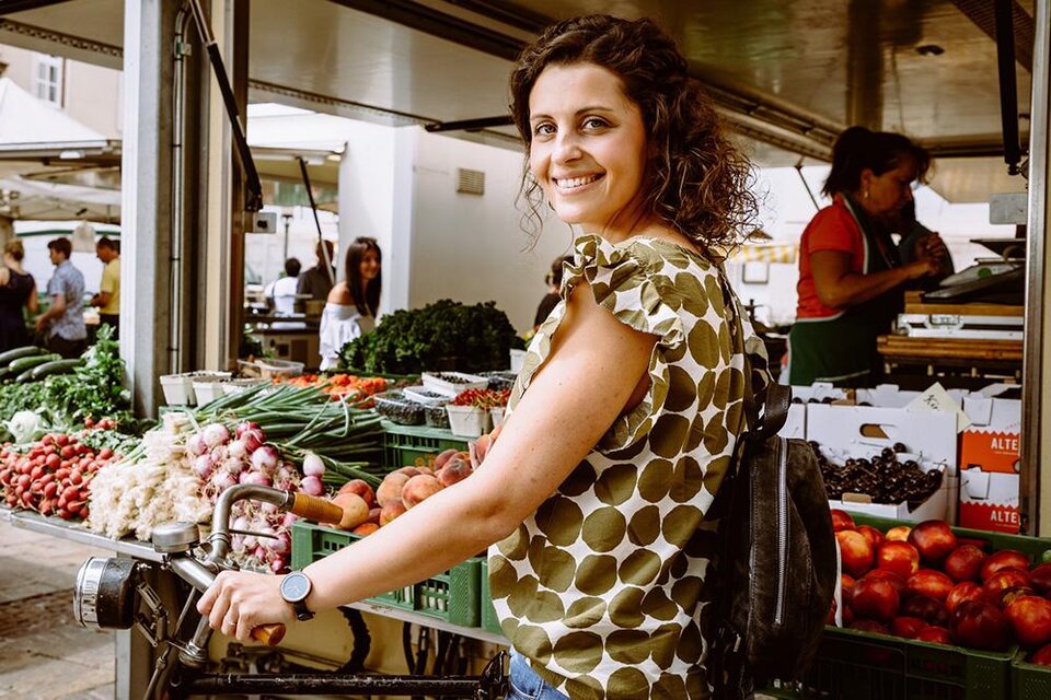 Eine Frau lächelt vor einem Marktstand mit frischem Obst und Gemüse. Im Hintergrund sind weitere Käufer und Verkäufer zu sehen. | © TVB Erzberg-Leoben