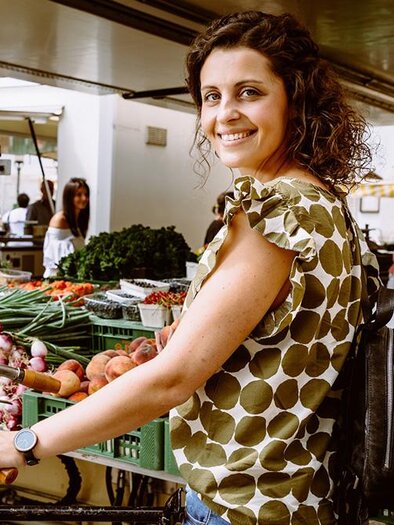 Eine Frau lächelt vor einem Marktstand mit frischem Obst und Gemüse. Im Hintergrund sind weitere Käufer und Verkäufer zu sehen. | © TVB Erzberg-Leoben