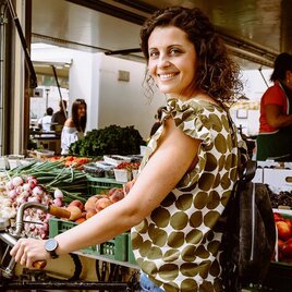 Eine fröhliche Frau steht auf einem Markt voller frischer Lebensmittel. Um sie herum sind Obst und Gemüse in bunten Kisten angeordnet. | © TVB Erzberg-Leoben