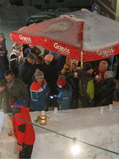 A bustling winter scene with many people gathering under a red umbrella. The ground is covered with snow and there are festive lights. | © Bergrettung