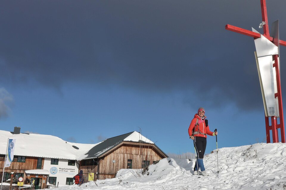Ein Schneegebiet mit einem Wanderer in roter Jacke, der auf einem schneebedeckten Hügel steht. Im Hintergrund ist ein Hinweis auf eine Berghütte und ein großes rot-weißes Kreuz sichtbar. | © TVB Erzberg-Leoben