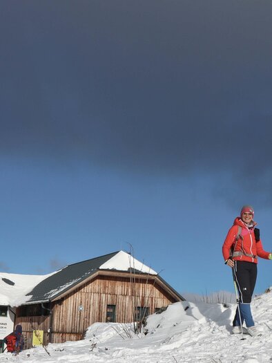 Ein Schneegebiet mit einem Wanderer in roter Jacke, der auf einem schneebedeckten Hügel steht. Im Hintergrund ist ein Hinweis auf eine Berghütte und ein großes rot-weißes Kreuz sichtbar. | © TVB Erzberg-Leoben