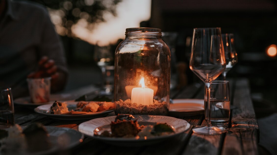Ein romantisches Abendessen im Freien mit Kerzenlicht und köstlichen Speisen. Der Tisch ist mit Gläsern und Tellern gedeckt, während es Dämmerung ist. | © TVB Südsteiermark – Lukas Elsneg