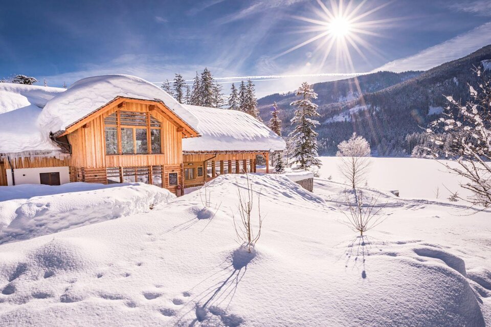 Ein gemütliches Holzhaus im Schnee mit einer strahlenden Sonne im Hintergrund. Die Landschaft ist winterlich und idyllisch.