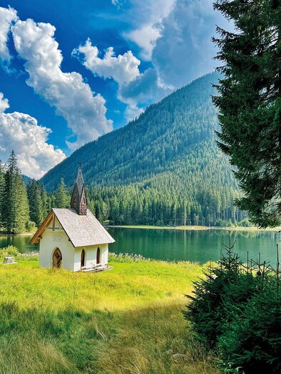 A picturesque landscape with a small house by the lake.  
Surrounded by tall mountains and dense forests under a blue sky with clouds. | © (c)KLOSTERWALD