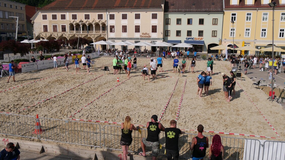 Ein Sandplatz mit vielen Menschen, die an einer Veranstaltung teilnehmen. Im Hintergrund sind bunte Gebäude und Restaurants zu sehen. | © Laura Schneller
