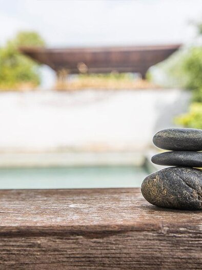 Three stacked stones on a wooden block. In the background, there is a calm pool and green trees. | © Bild: Freepik