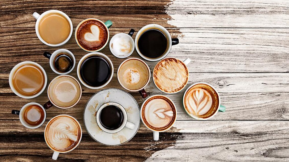 Eine Vielzahl von Kaffeetassen auf einem Holztisch. Einige Tassen haben kunstvolle Milchschaummuster. | © Bild: freepik