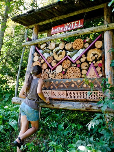 An insect hotel made of natural materials stands amidst trees and plants. A person is curiously observing it. | © Holzmuseum