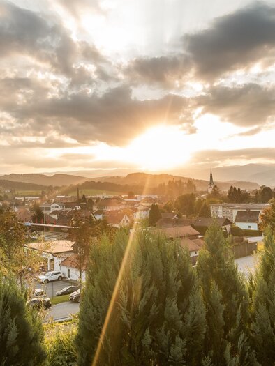 Ein malerischer Sonnenuntergang über einer kleinen Stadt, umgeben von Hügeln. Die Wolken reflektieren das warmes Licht der Sonne. | © Lipizzanerheimat-DieAbbilderei