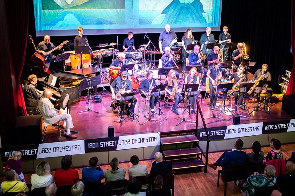 Ein großes Jazzorchester spielt auf einer Bühne, umgeben von Instrumenten. Die Zuschauer sitzen im Publikum und genießen die Darbietung. | © Iron Street Jazz Orchestra