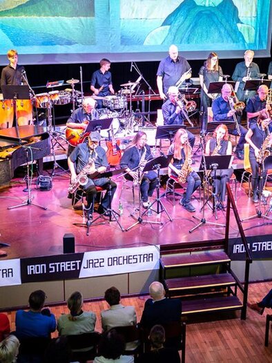 A large jazz orchestra plays on a stage, surrounded by instruments. The audience sits in the crowd and enjoys the performance. | © Iron Street Jazz Orchestra