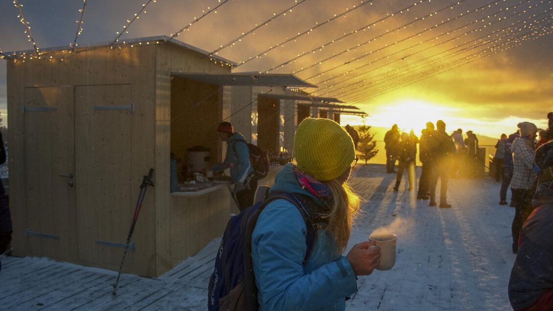 Eine Person steht in einer schneebedeckten Landschaft und hält eine Tasse in der Hand. Im Hintergrund ist ein schöner Sonnenuntergang zu sehen, begleitet von festlichen Lichtern. | © Bergbahnen Stuhleck