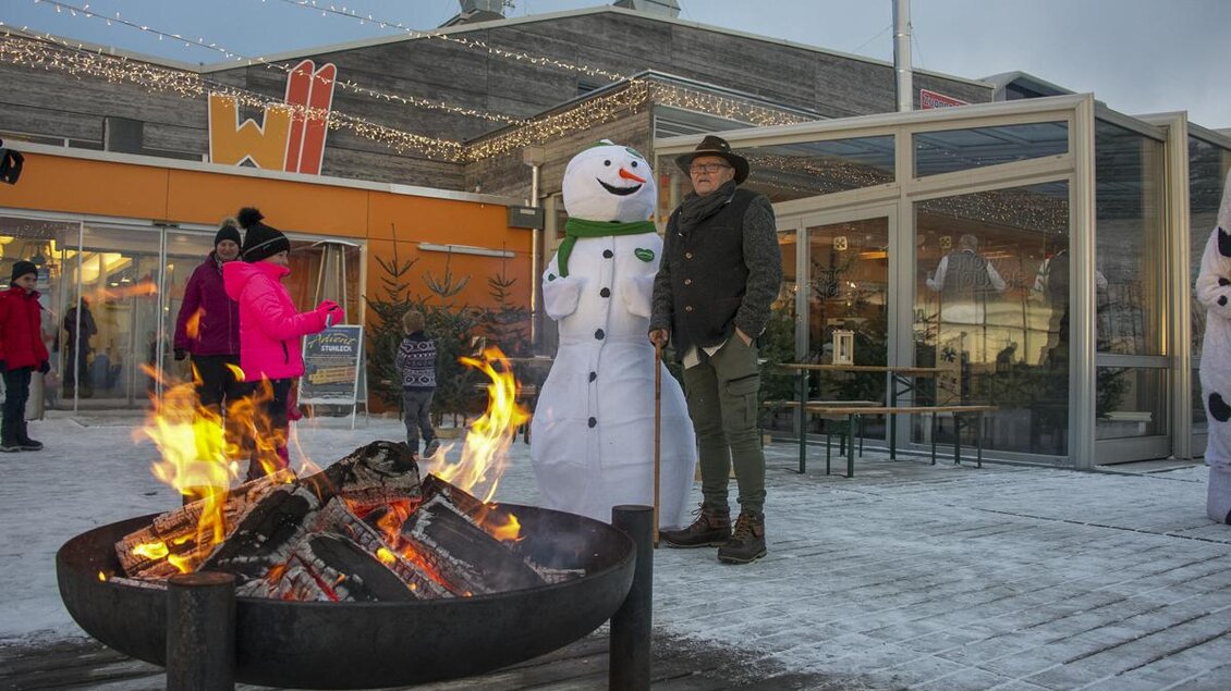 Ein gemütliches Winterambiente mit einem Feuer und einem Schneemann. Eine Person steht auf einem Holzdeck vor einem Gebäude, umgeben von winterlicher Dekoration. | © Bergbahnen Stuhleck