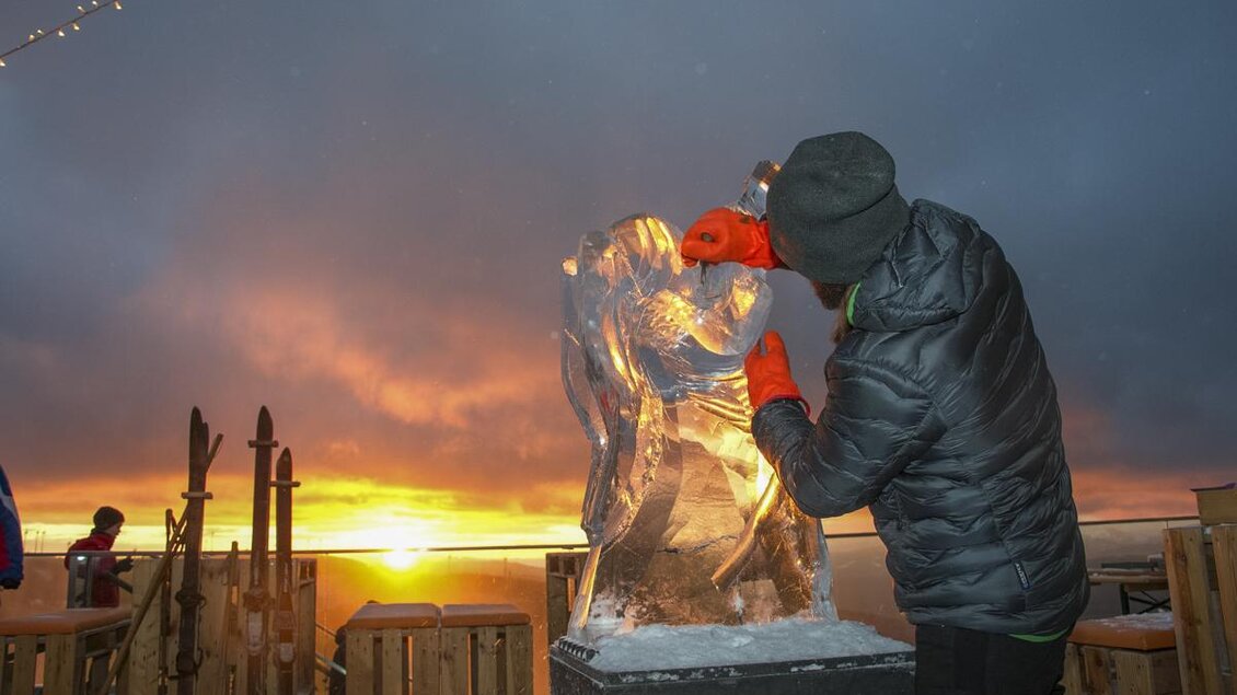 Ein Künstler arbeitet an einer Eisfigur bei Sonnenuntergang. Die dramatische Beleuchtung verstärkt die Schönheit der Skulptur und der Umgebung. | © Bergbahnen Stuhleck
