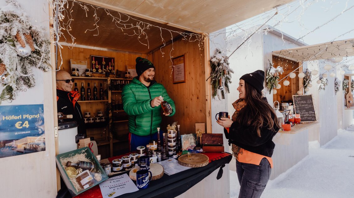 Ein stimmungsvolles Marktstand mit einem Verkäufer und einer Kundin. Der Stand bietet verschiedene lokale Produkte und ist winterlich dekoriert. | © nicoleseiser.at