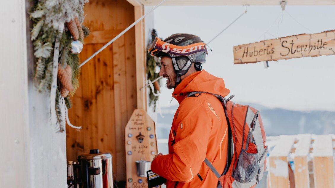 Ein Skifahrer in einem orangefarbenen Anzug steht an einem Verkaufsstand in den Bergen. Im Hintergrund sind winterliche Dekorationen und eine Holztafel mit dem Namen „Hubertus Sternhütte“ zu sehen. | © nicoleseiser.at