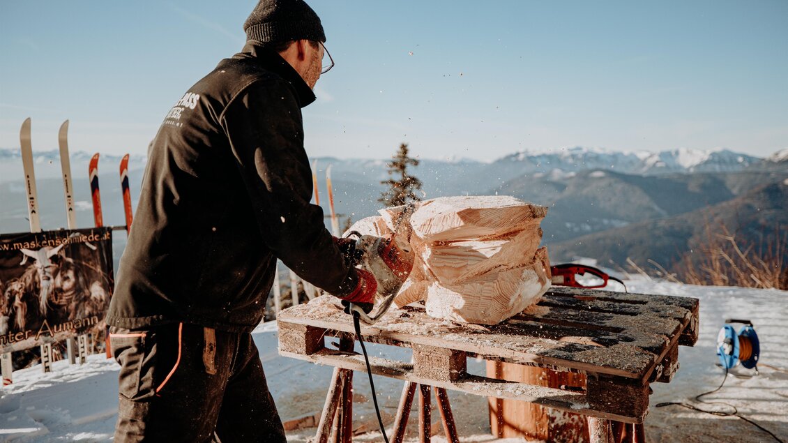 Ein Handwerker schnitzt Holzskulpturen im Schneegebirge. Im Hintergrund sind Berge und Skier zu sehen. | © nicoleseiser.at