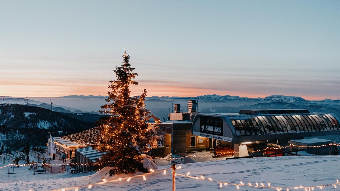 Eine Winterlandschaft mit einem gemütlichen Gebäude und einem beleuchteten Weihnachtsbaum. Der Himmel ist in sanften Farben des Sonnenuntergangs gefärbt. | © nicoleseiser.at