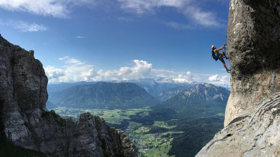 Ein Kletterer, der an einer steilen Felswand hochsteigt. Im Hintergrund sind beeindruckende Berge und ein blaues Himmel zu sehen. | © Loser Bergbahnen Altaussee_Markus Raich