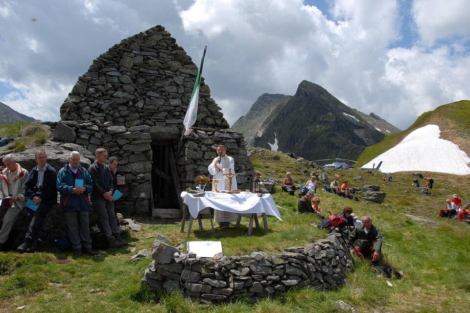 Eine Gruppe von Menschen versammelt sich im Freien vor einer alten Steinruine in den Bergen. Im Vordergrund befindet sich ein Tisch mit einer Decke, auf dem möglicherweise eine Zeremonie oder Feier stattfindet.