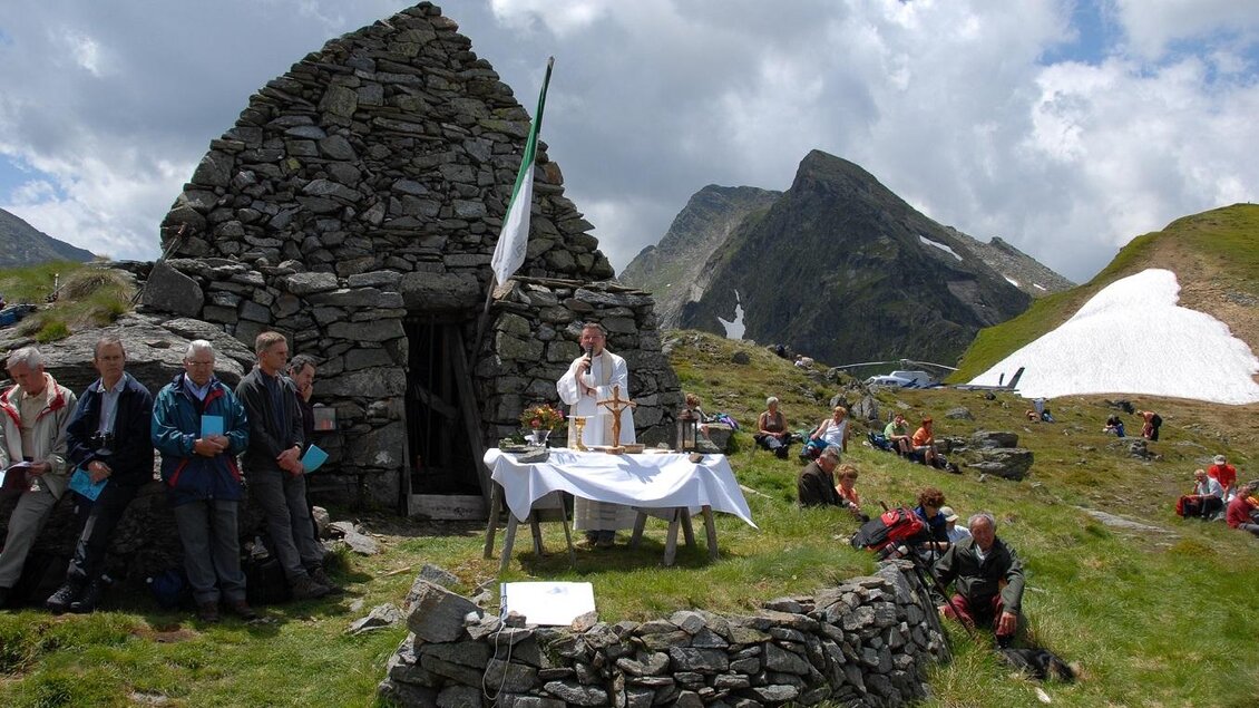 Eine Gruppe von Menschen versammelt sich im Freien vor einer alten Steinruine in den Bergen. Im Vordergrund befindet sich ein Tisch mit einer Decke, auf dem möglicherweise eine Zeremonie oder Feier stattfindet.