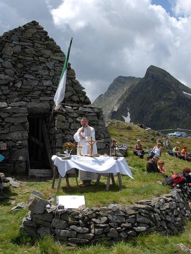 Eine Gruppe von Menschen versammelt sich im Freien vor einer alten Steinruine in den Bergen. Im Vordergrund befindet sich ein Tisch mit einer Decke, auf dem möglicherweise eine Zeremonie oder Feier stattfindet.