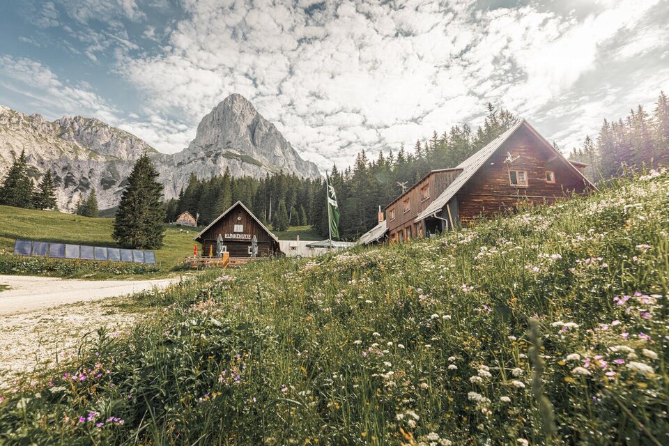 Eine idyllische Berglandschaft mit traditionellen Hütten und einer grünen Wiese voller Blumen. Im Hintergrund erheben sich majestätische Berge unter einem bewölkten Himmel. | © Thomas Sattler