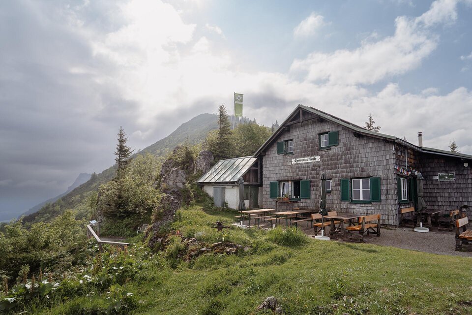 Eine malerische Berghütte umgeben von grünen Wiesen und Bäumen. Im Hintergrund sind hohe Berge und ein bewölkter Himmel zu sehen. | © TV Gesäuse