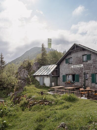 Eine malerische Berghütte umgeben von grünen Wiesen und Bäumen. Im Hintergrund sind hohe Berge und ein bewölkter Himmel zu sehen. | © TV Gesäuse