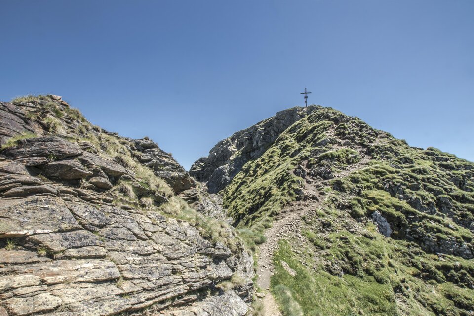 Ein malerischer Bergpfad führt zu einem Gipfel mit einem Kreuz. Die Landschaft ist grün und von Felsen umgeben unter klarem blauem Himmel. | © Gerhard Pilz