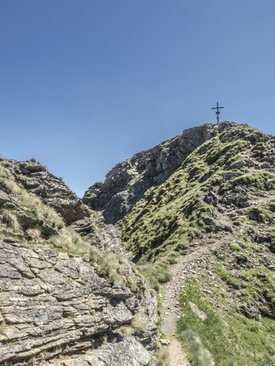 Ein malerischer Bergpfad führt zu einem Gipfel mit einem Kreuz. Die Landschaft ist grün und von Felsen umgeben unter klarem blauem Himmel. | © Gerhard Pilz