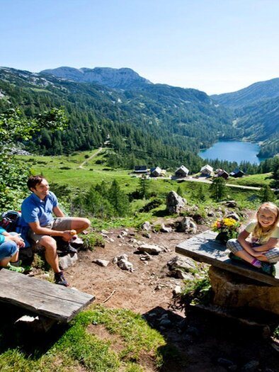 A family is sitting on a bench in the mountains, enjoying the view of a clear lake. In the background, there are green forests and majestic mountains visible. | © TVB Ausseerland_Tom Lamm