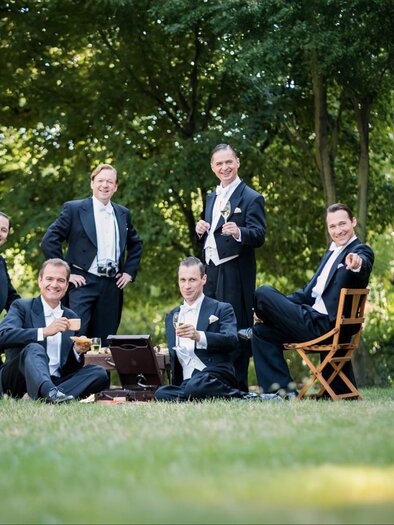 Sechs gut gekleidete Männer sitzen in einem grünen Park. Sie genießen Drinks und sind fröhlich zusammen. | © Oliver Betke