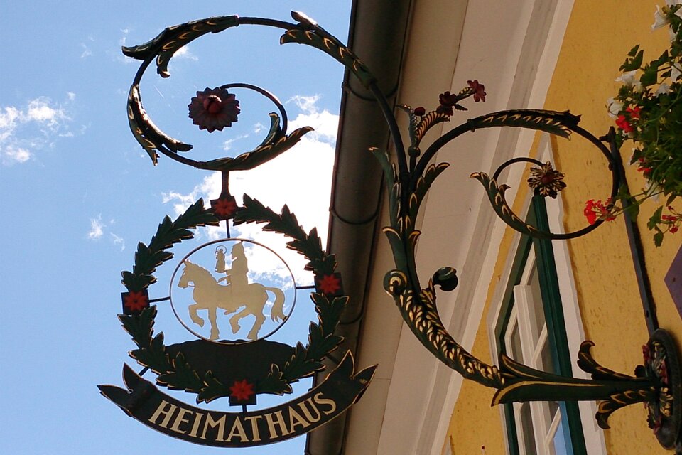 Ein dekoratives Schild mit dem Schriftzug "HEIMATHAUS". Es zeigt ein Wappen mit einem Pferd und Blumen. | © Fred Lindmoser
