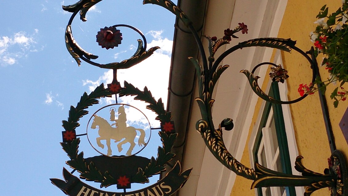 Ein dekoratives Schild mit dem Schriftzug "HEIMATHAUS". Es zeigt ein Wappen mit einem Pferd und Blumen. | © Fred Lindmoser