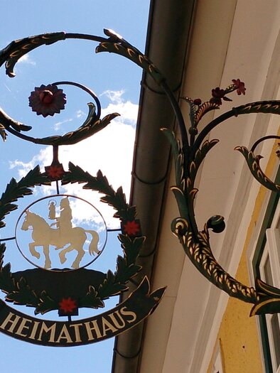 Ein dekoratives Schild mit dem Schriftzug "HEIMATHAUS". Es zeigt ein Wappen mit einem Pferd und Blumen. | © Fred Lindmoser
