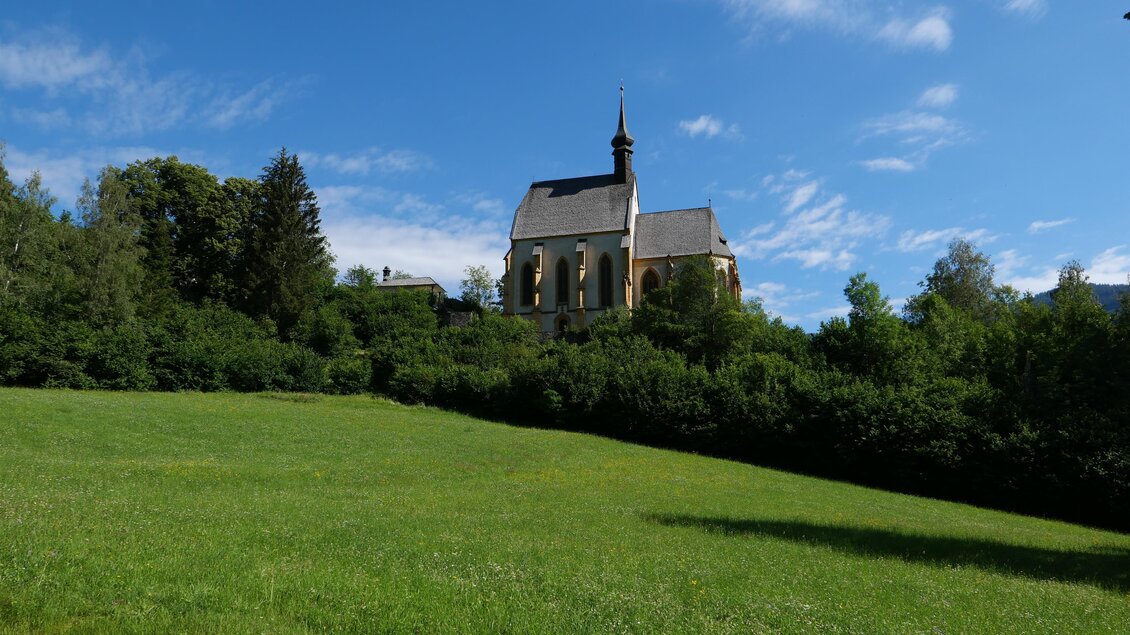 Eine alte Kirche steht auf einem grünen Hügel, umgeben von Bäumen. Der Himmel ist blau mit einigen Wolken.