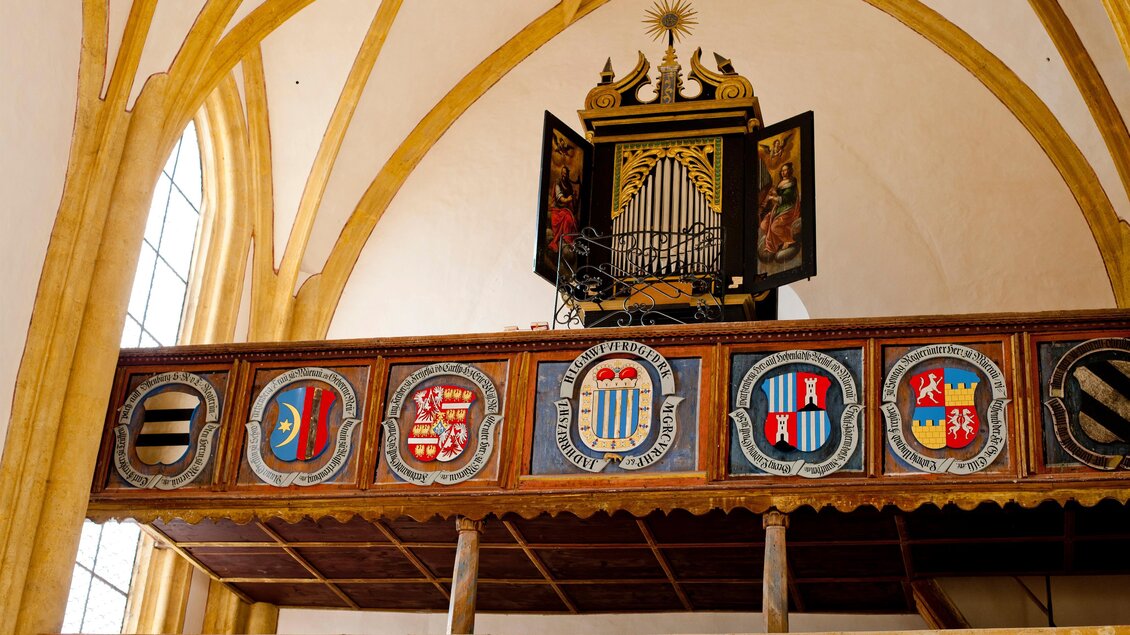 Eine kunstvolle Orgel im Innenraum einer Kirche, umgeben von detaillierten Wappen. Die hohen, gewölbten Decken verleihen dem Raum eine beeindruckende Atmosphäre.