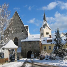 Ein malerisches Winterdorf mit schneebedeckten Dächern und einer Kirche mit Turm. Die Landschaft wirkt ruhig und einladend.