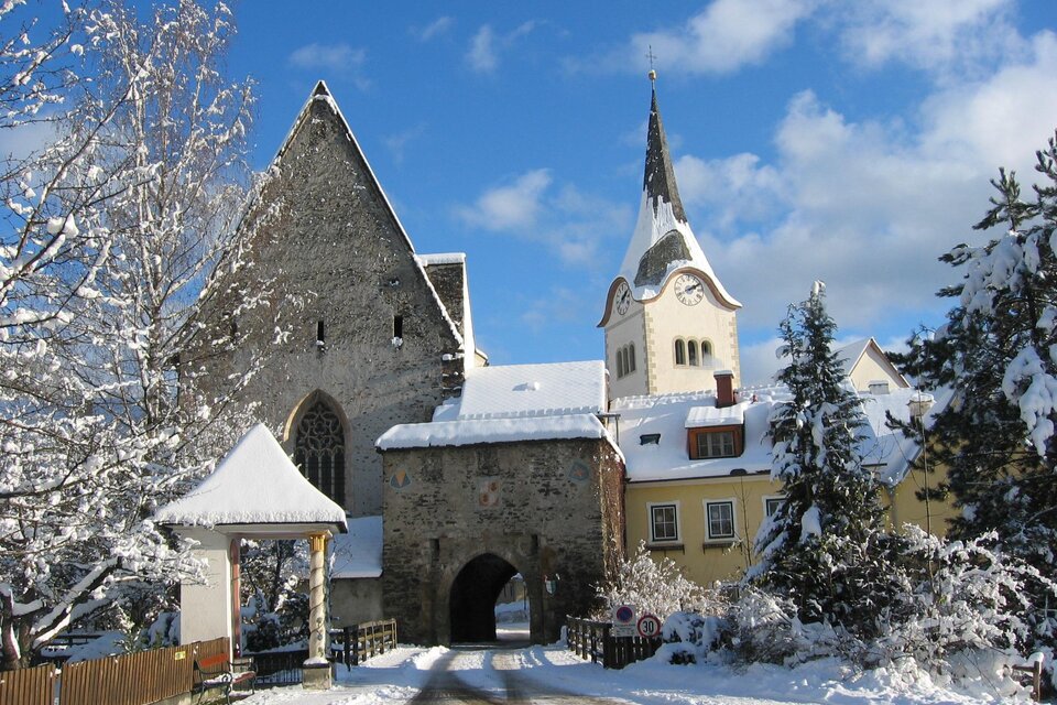 Ein malerisches Winterdorf mit schneebedeckten Dächern und einer Kirche mit Turm. Die Landschaft wirkt ruhig und einladend.