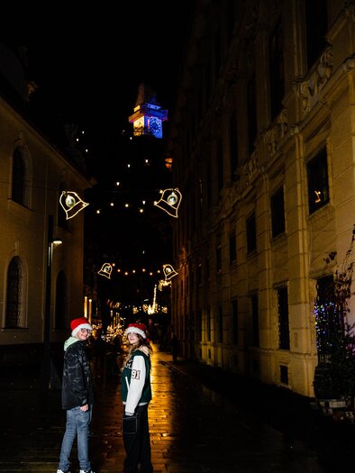 Two people wearing Christmas hats stand in a festively lit street. The path is wet and shiny, while lights sparkle on the buildings. | © (c) Region Graz - Mias Photoart