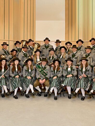 A large group of people in traditional costumes is standing together in a bright room. Some are holding musical instruments and have flowers or honors in their hands. | © Kerstin Zmugg