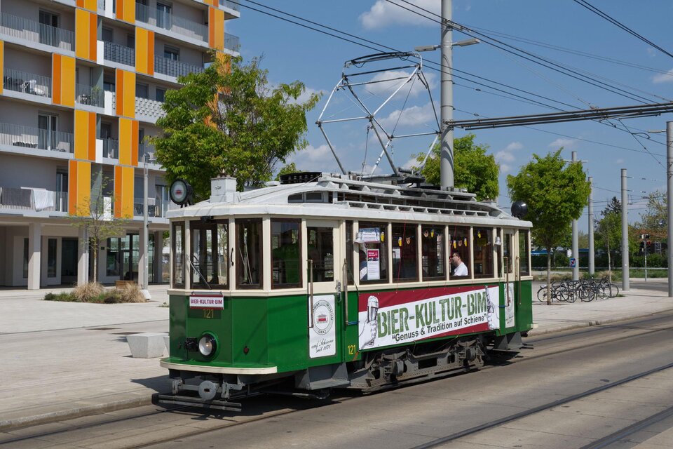Eine historische Straßenbahn fährt auf einer urbanen Straße. Im Hintergrund sind moderne Gebäude und Bäume zu sehen. | © (C)Tramway Museum Graz