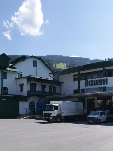 Eine Brauerei mit einem großen Gebäude und einem klaren Himmel. Es sind mehrere Fahrzeuge und Menschen vor der Brauerei zu sehen. | © Gerhard Pilz