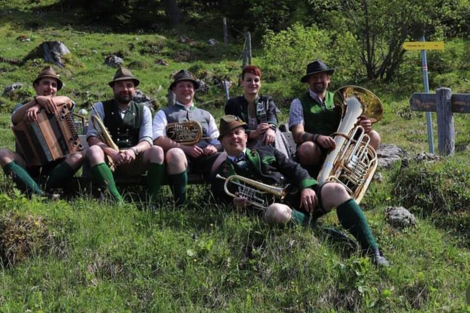 Die Musikanten von D'Röthelstoana in traditioneller Tracht  mit ihren Instrumenten der klassischen Volksmusik sitzen auf einer sommerlichen Wiese im Ausseerland.  | © Das Schutzhaus / D'Röthelstoana
